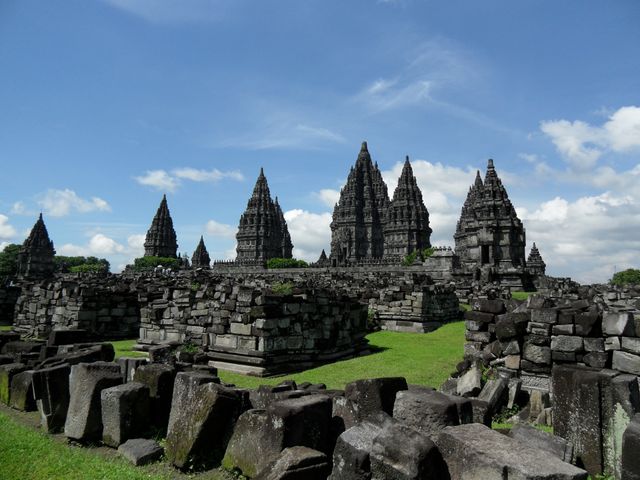 Prambanan temple complex shows ancient Hindu architecture with tall stone structures set against a blue sky. Green grass surrounds temple area, presenting a serene and captivating environment. Ideal for use in travel publications, cultural and historical brochures, educational materials on ancient architecture, and travel blogs highlighting Indonesian landmarks.