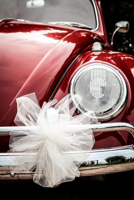 Close-up view of a red vintage car decorated with white tulle bow in front of headlight, often used for wedding transportation. Great for wedding invitations, blogs, or event planning resources highlighting classic vehicle charm and bridal motifs.
