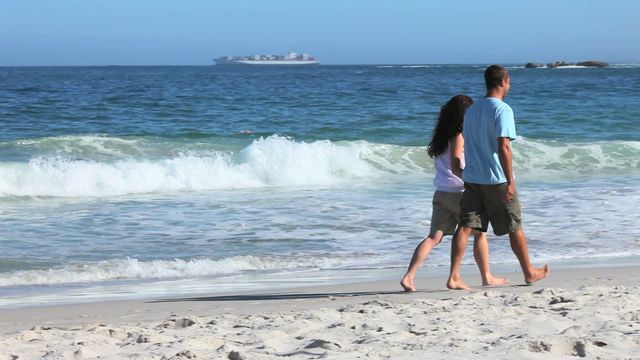 A couple walks on a sandy beach with gentle waves in the background. They enjoy a sunny day, casting long shadows on the sand. Ideal for use in travel brochures, romantic retreat advertisements, or lifestyle blogs focusing on relaxation and summer activities.