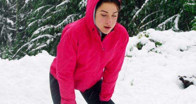 Woman taking a break from her run in a snowy forest. She is resting with hands on knees while wearing a bright red jacket. Can be used for articles on winter exercise tips, fitness motivation, outdoor activities, and healthy lifestyle during cold seasons.