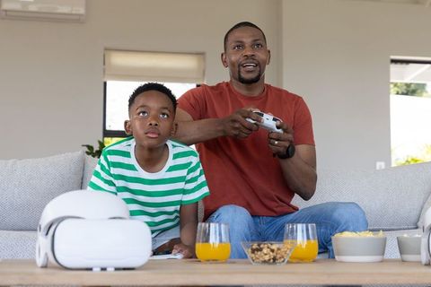African american father and son playing video games on couch with vr headset and snacks