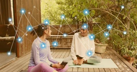 Women talking on porch yoga mats holding smartphone with network overlay for wellness