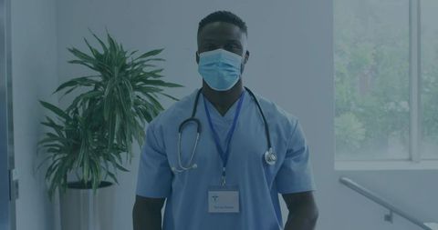 Masked Male Clinician Wearing Blue Scrubs, Stethoscope and ID Badge in Clinic Corridor
