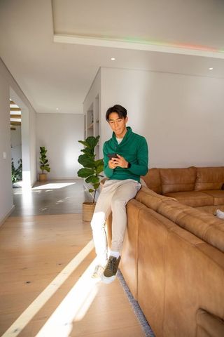 Twentysomething asian man sitting on leather sectional using smartphone in sunlit modern living room