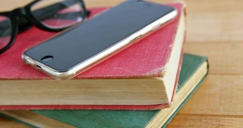 Smartphone resting on worn red book with reading glasses on wooden table closeup vintage