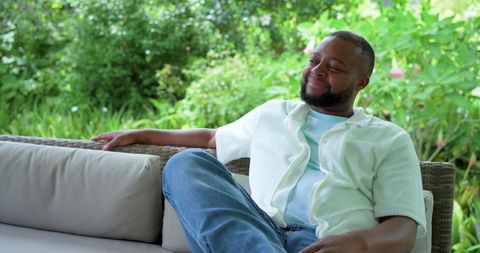 Relaxing and smiling on wicker outdoor sofa in sunny garden wearing casual shirt and jeans
