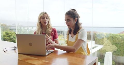 Young women collaborating on laptop at sunlit glass-walled table overlooking garden
