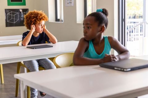 Classmates sitting apart in bright classroom looking thoughtful with digital devices