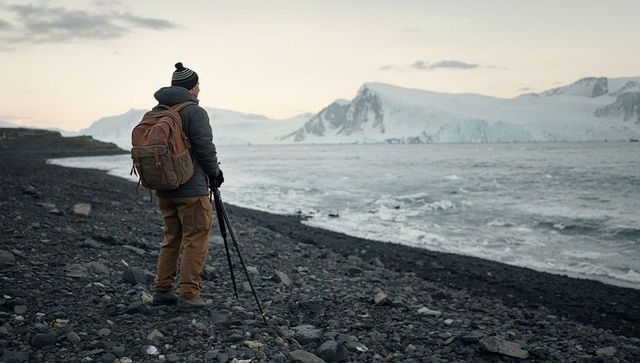 Positioning tripod on rocky arctic coast, man overlooking glaciers and rough sea at dusk