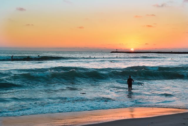 Man holding surfboard and looking at ocean waves while sun sets. Ideal for themes related to summer vacations, surfing adventures, beach holidays, and outdoor activities. Suitable for travel blogs, adventure promotions, and surf school advertisements.