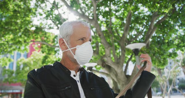 Elderly man wearing a protective face mask outdoors in an urban setting with trees in the background. This can be used to illustrate concepts related to health and safety, public health measures, mature population during a pandemic, or outdoor activities during health crises.