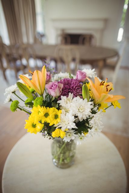 Colorful flower bouquet in a glass vase placed on a round table in a home interior. The arrangement includes lilies, daisies, chrysanthemums, and roses, creating a vibrant and fresh look. Ideal for use in articles or advertisements related to home decor, interior design, floral arrangements, and lifestyle blogs.