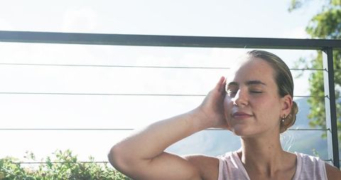 Sunlit woman resting on balcony, leaning against cable railing with eyes closed, serene