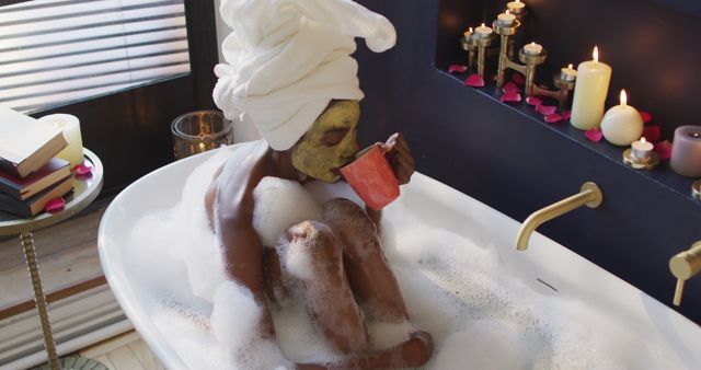 African American woman with towel on head and face mask relaxing in a foam-filled bathtub, enjoying a hot drink. Candles and rose petals adding to serene atmosphere. Perfect for concepts related to self-care, wellness, relaxation, home spa routines, and pampering time.