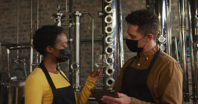 Two minority brewery workers are seen examining a glass of beer within an industrial brewery setting. Both are wearing black masks and aprons, adhering to safety protocols. This image can be used to depict workplace safety, teamwork, industrial processes, and the brewing industry.