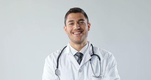 A young male doctor wearing a white coat and a stethoscope around his neck is standing while smiling confidently. This image is perfect for promoting healthcare services, medical clinics, hospitals, or educational materials related to medicine and health professions.