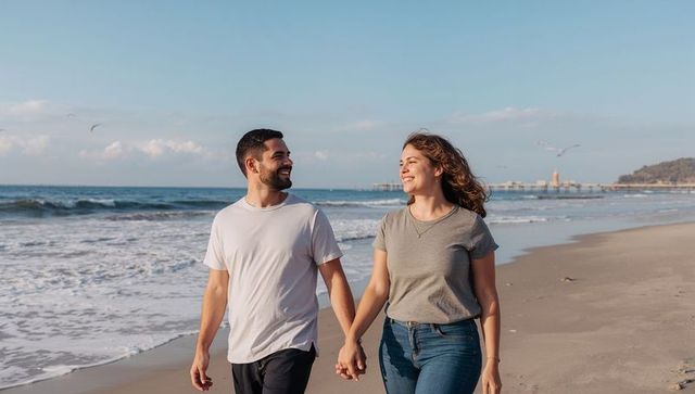 Couple walking hand in hand on sandy beach at golden hour smiling by ocean pier