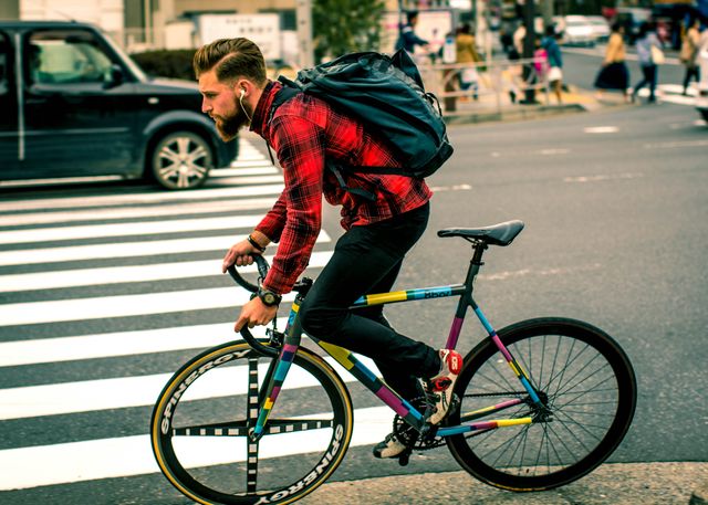 Bearded man riding a colorful bicycle in an urban area, wearing a red plaid shirt and black pants, with a large backpack over his shoulders. Use this image for themes related to daily commutes, urban exploration, youth culture, active transport, or modern lifestyle.