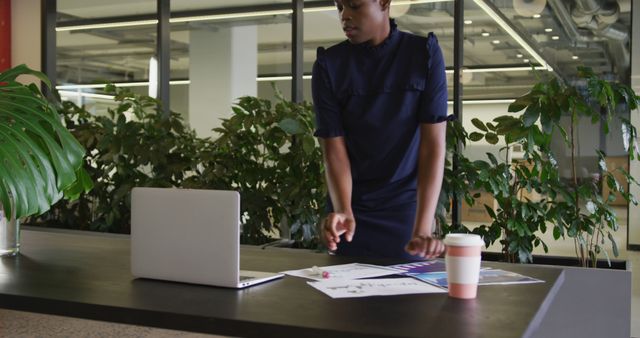 Businesswoman stands at desk reviewing documents in a contemporary office space. Ideal for depicting professional work environments, modern office settings, project management, or corporate lifestyle themes.