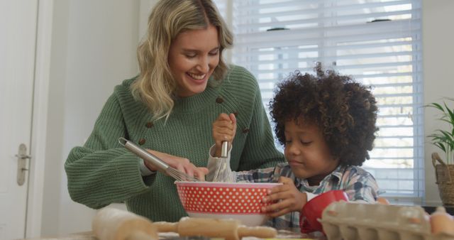 Mother and child enjoy quality time baking together in a kitchen. They are whisking ingredients in a red polka dot bowl, smiling and engaging. Ideal for content related to family activities, parenting, cooking classes, and home bonding moments.