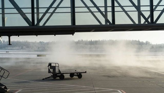 Empty baggage cart on frosty airport apron under glass jetway with morning mist