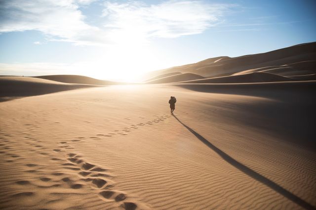Image shows individual walking on vast desert sand dunes with sun setting in background, casting long shadows. Footprints trail behind person, emphasizing sense of adventure and exploration. Ideal for use in travel blogs, adventure magazines, trekking advertisements, and publications focusing on solitude or nature.