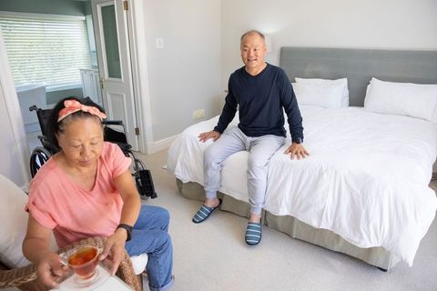 Senior asian couple sharing tea in cozy bedroom, man sitting on bed smiling, wheelchair nearby