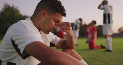 Young soccer player sitting, pinching nose after match loss at dusk on grassy field