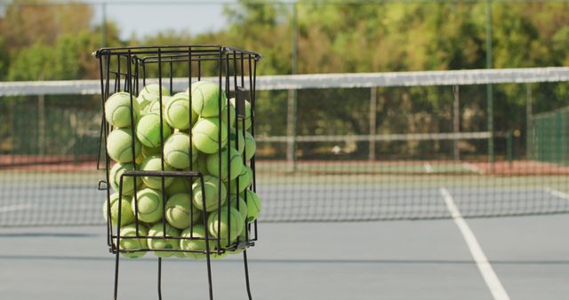 Tennis balls kept in a wire basket on an outdoor tennis court. Suitable for articles about tennis training, sports equipment, or fitness activities. Ideal for websites, blogs, and advertisements promoting outdoor sports, training sessions, or tennis facilities.