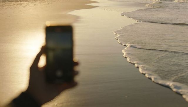Holding smartphone capturing golden hour shoreline with foamy waves and silhouette