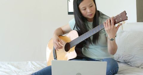 Asian woman practicing acoustic guitar on bed at home while following online lesson on laptop