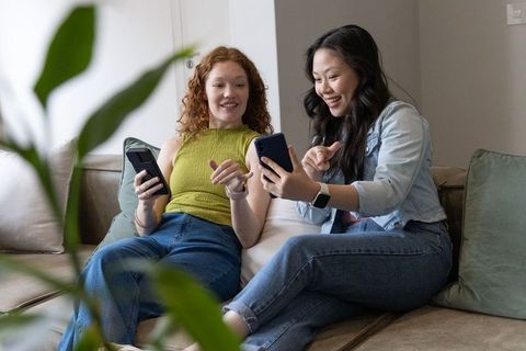 Two friends sharing smartphone on sofa, laughing and scrolling, casual home lifestyle