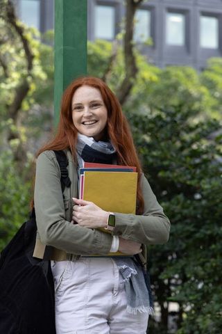 Redhead student leaning on green pole holding folders and smiling on campus