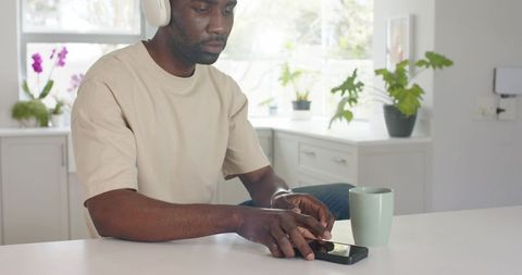 Young man tapping smartphone wearing over-ear headphones at bright kitchen counter with green mug