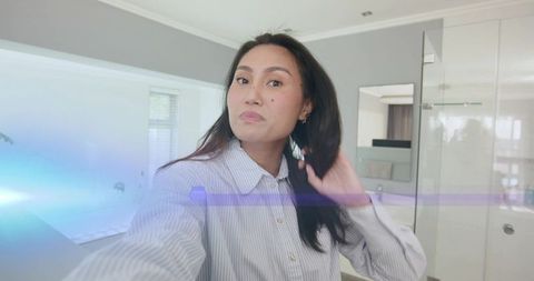 Asian woman taking mirror selfie while adjusting hair wearing striped shirt in bathroom