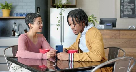 Multicultural couple sharing smartphone and coffee at glossy table in modern cafe kitchen