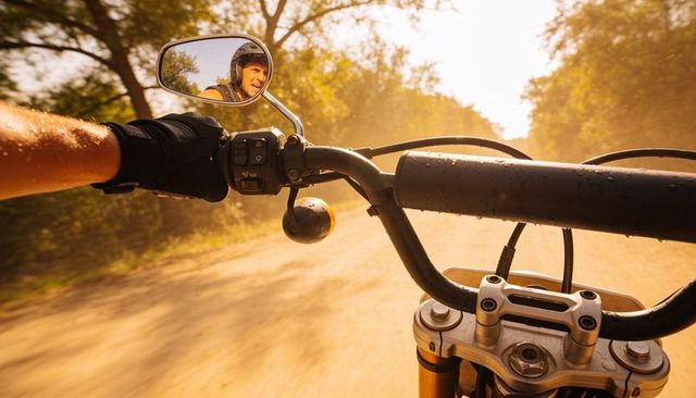 Riding motorcycle on dusty sunlit country road with gloved hand on throttle and mirror
