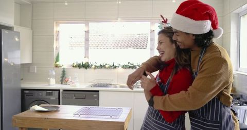 Couple hugging in festive kitchen wearing santa hat and reindeer antlers, holiday cheer