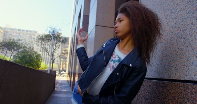 Young woman with curly hair striking a confident pose in modern urban area. Wears stylish leather jacket and casual outfit, sunlight highlighting her features. Ideal for use in fashion editorials, lifestyle blogs, street style photography, and modern urban living promotions.