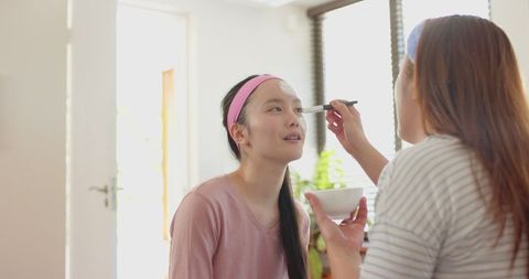 Mother and Daughter Applying Facial Mask Near Window and Bonding at Home