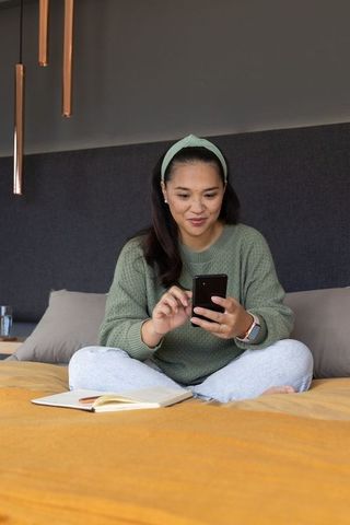 Asian woman sitting cross-legged on bed using smartphone and notebook in cozy minimalist bedroom
