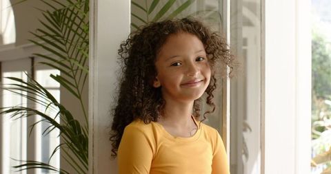 Smiling girl leaning against doorway among sunlit potted palms in warm home interior