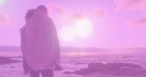 Couple wrapped in blanket embracing watching pastel pink-purple sunset over rocky shoreline