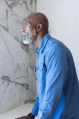 Senior african man leaning on counter reflecting in sunlit marble bathroom wearing denim