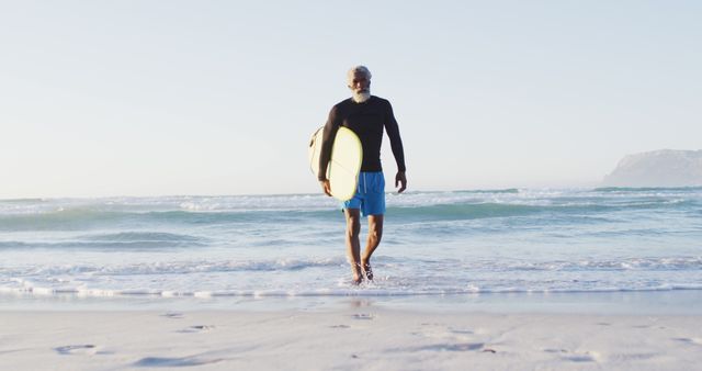 Senior african american man walking with surfboard on sunny beach. healthy and active time beach holiday.