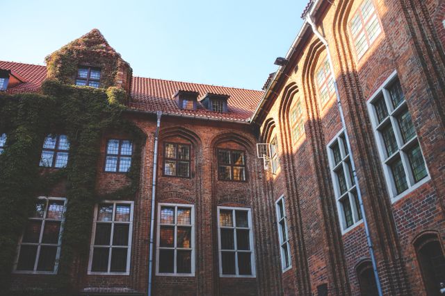 The image presents a historic building with red brick walls, multiple windows, and ivy growing on the facade. The medieval architectural style features tall arched windows and a tiled rooftop. Sunlight illuminates part of the building, casting shadows that add a dramatic effect. Ideal for themes like European travel, historical sites, architecture, urban exploration, and vintage aesthetics.