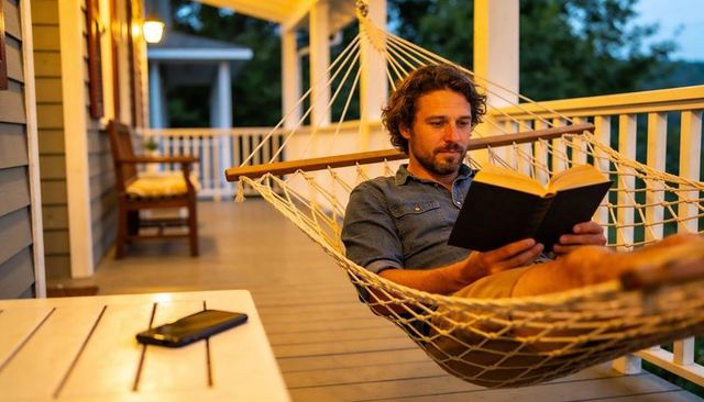 Man relaxing in hammock reading hardcover book on cozy evening porch with smartphone