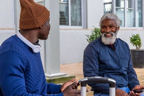 African american friends sitting outdoors smiling while talking with luggage and phones