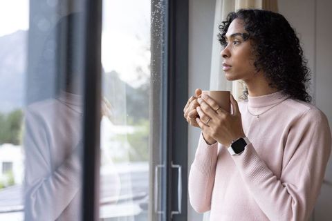Non-binary african american holding mug and gazing through rain-speckled window in pink sweater