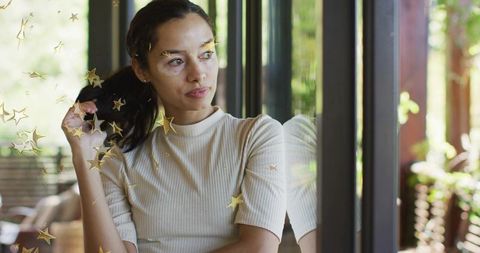 Contemplative woman gazing through window with golden star reflections, sunlit interior